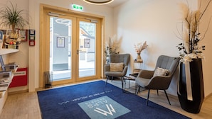 A reception area with a blue carpet, two grey chairs, a tall black vase with white flowers, and a wooden door with glass panels.