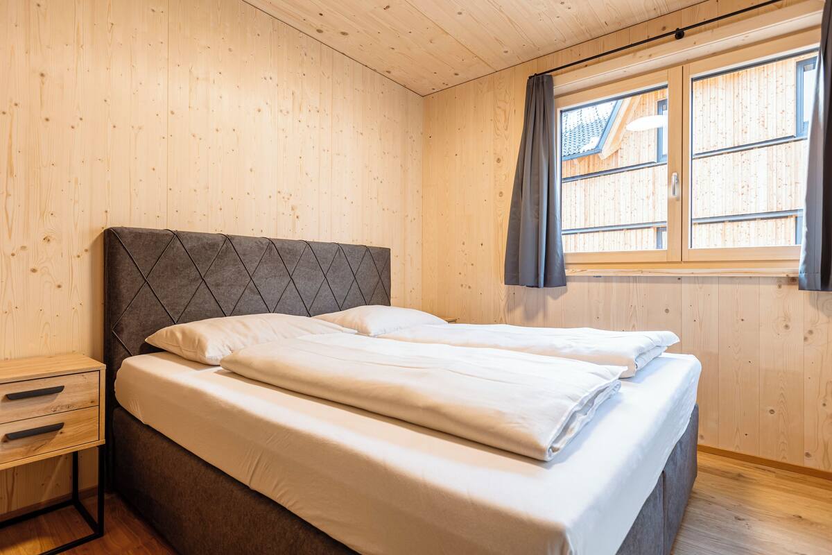 Une chambre à coucher avec un lit, des murs en bois, une fenêtre avec des rideaux et une table de chevet avec des tiroirs.