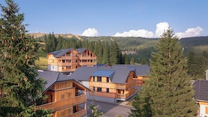 A multi-story wooden lodge with balconies, surrounded by dense forest and a mountainous backdrop.