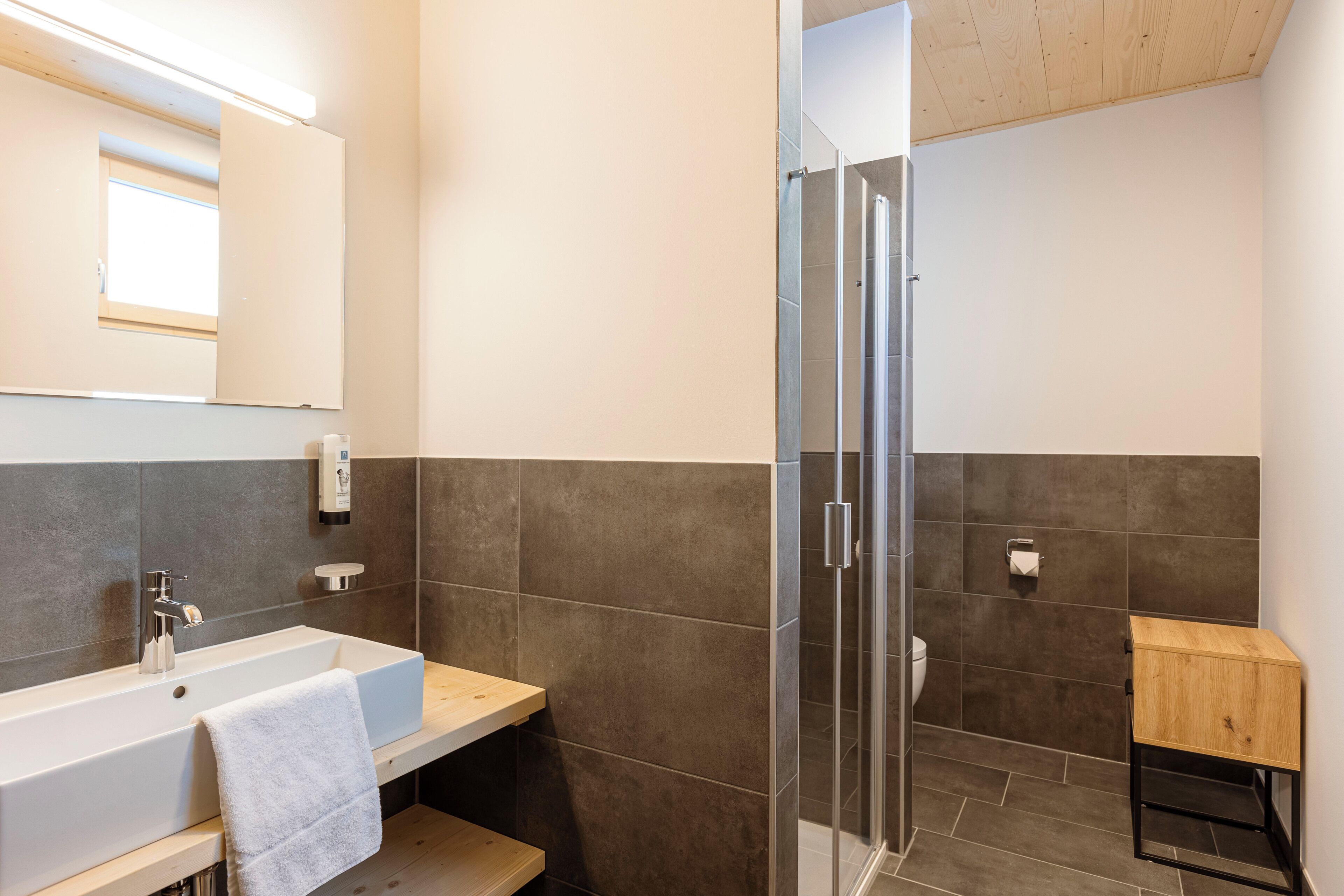 A modern bathroom with a large sink, a mirror, a shower enclosure, and a small wooden cabinet.