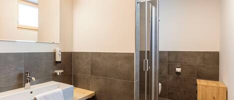 A modern bathroom with a large sink, a mirror, a shower enclosure, and a small wooden cabinet.
