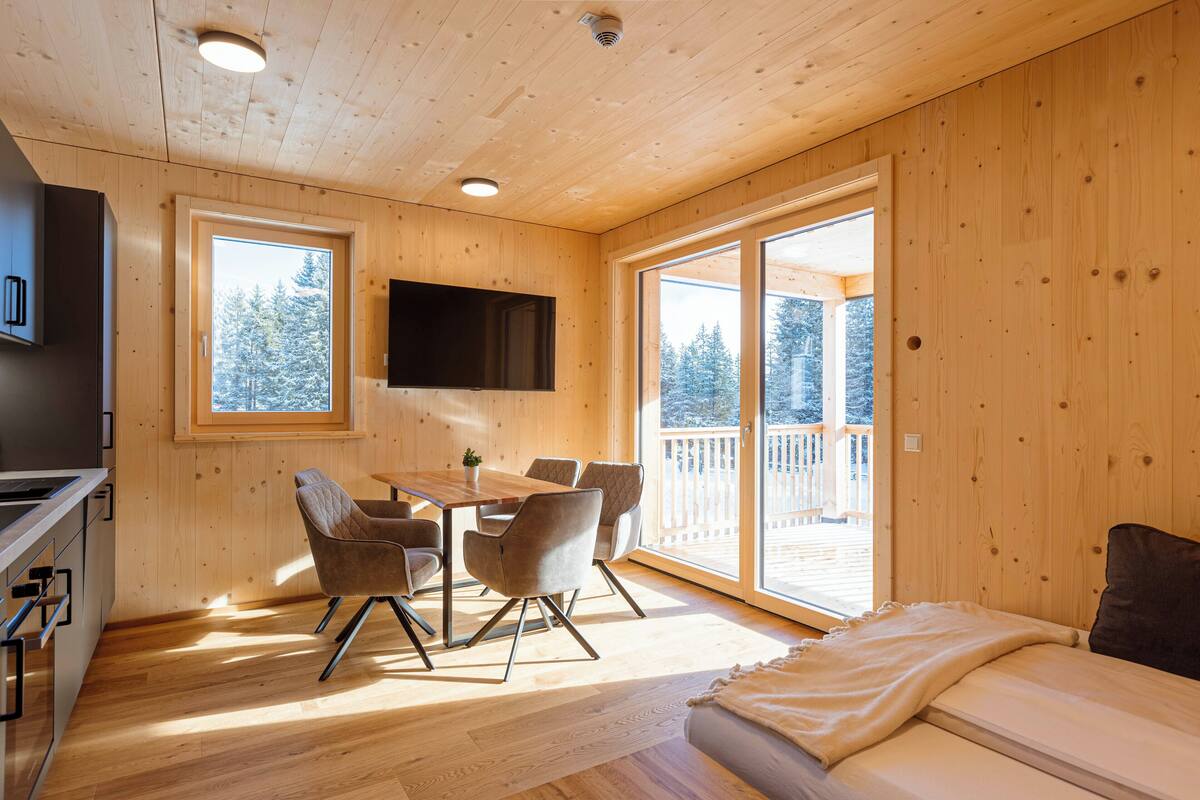 L’intérieur d’un chalet en bois comprend un coin repas, un canapé, un téléviseur fixé au mur et un balcon offrant une vue sur les arbres.