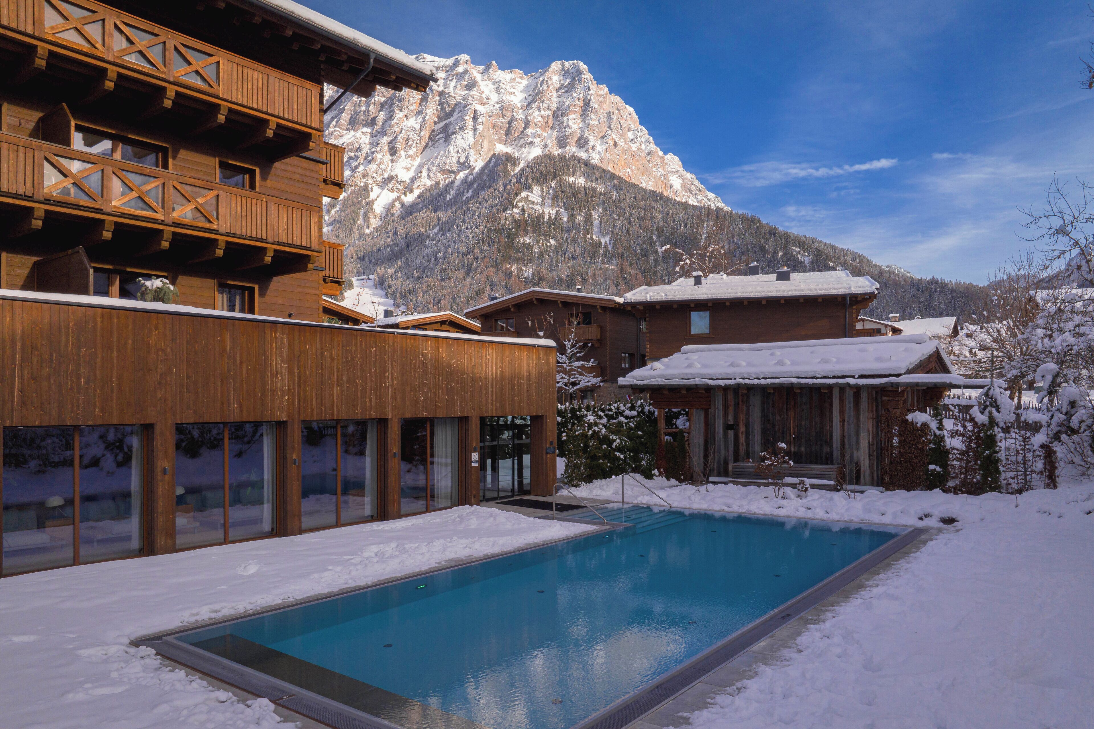 Un edificio de madera con piscina frente a él, rodeado de nieve y montañas al fondo.