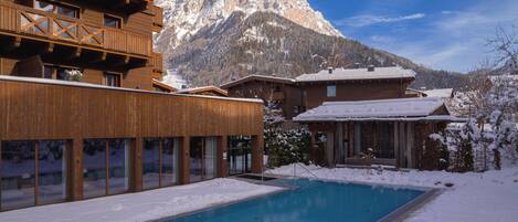 A wooden building with a pool in front, surrounded by snow and mountains in the background.