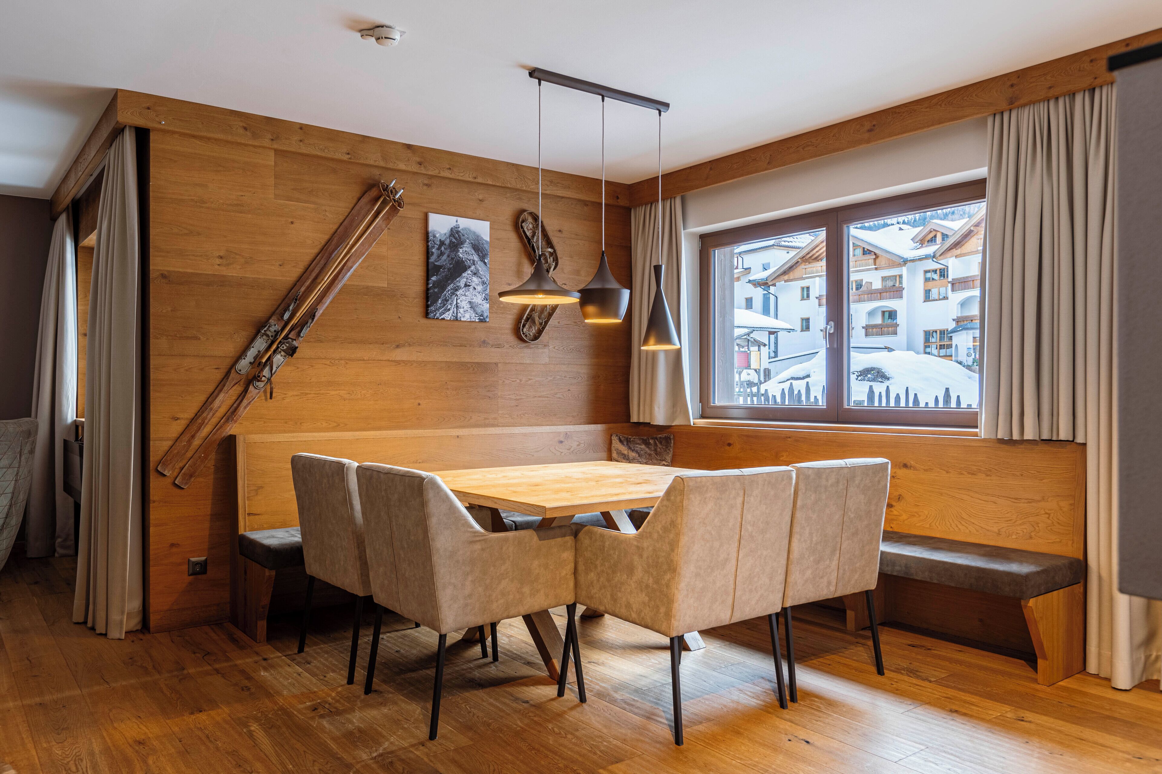 Una stanza con rivestimento in legno, un tavolo da pranzo in legno, quattro sedie, una panca, un portasci e una finestra con vista sugli edifici.