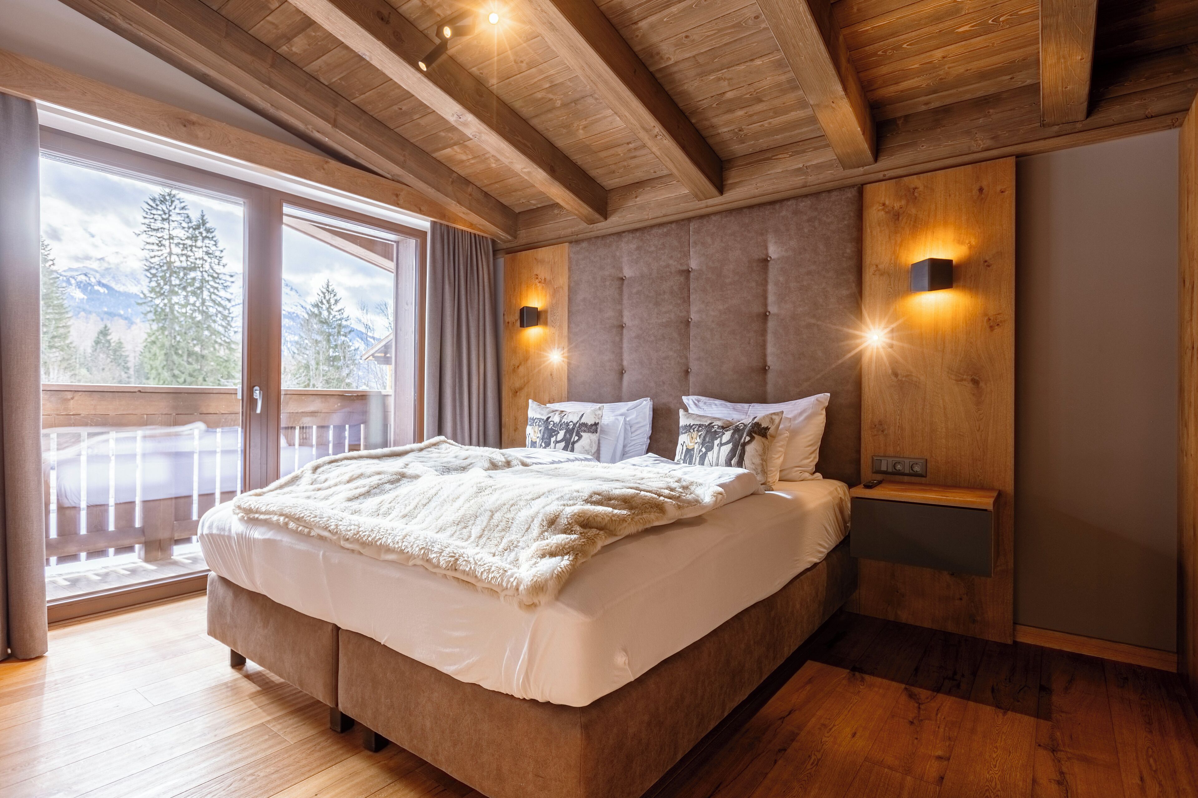 Une chambre dotée d’un grand lit, de poutres apparentes en bois au plafond, d’une porte-fenêtre coulissante donnant sur un balcon et de deux lampes fixées au mur.