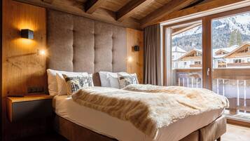 A bedroom with a large bed, wooden headboard, and a view of mountain houses through a glass door.