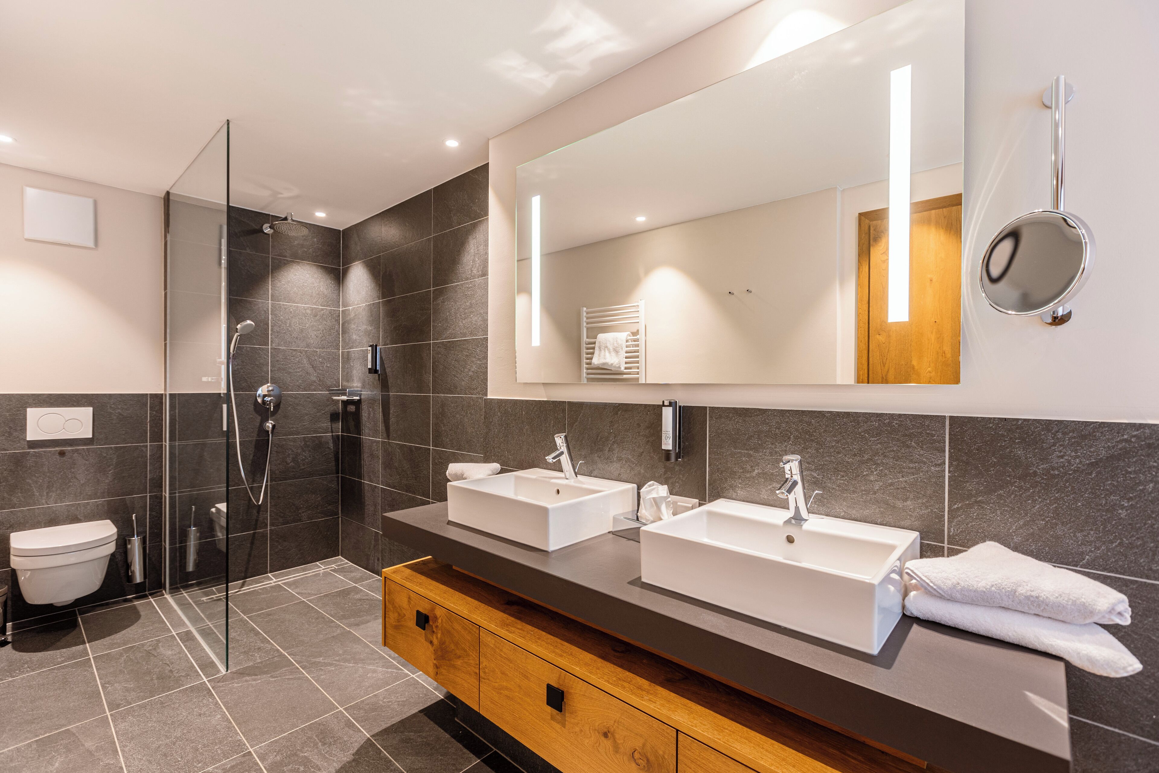 Ein modernes Badezimmer mit zwei Waschbecken, einer Duschkabine aus Glas und einem Holz-Waschtisch.