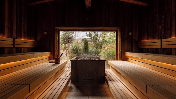 A sauna with wooden benches and a central hot tub, surrounded by a wooden structure with a view of trees and a building in the background. - Bergresort Zugspitze Ehrwald by ALPS RESORTS (Ehrwald)