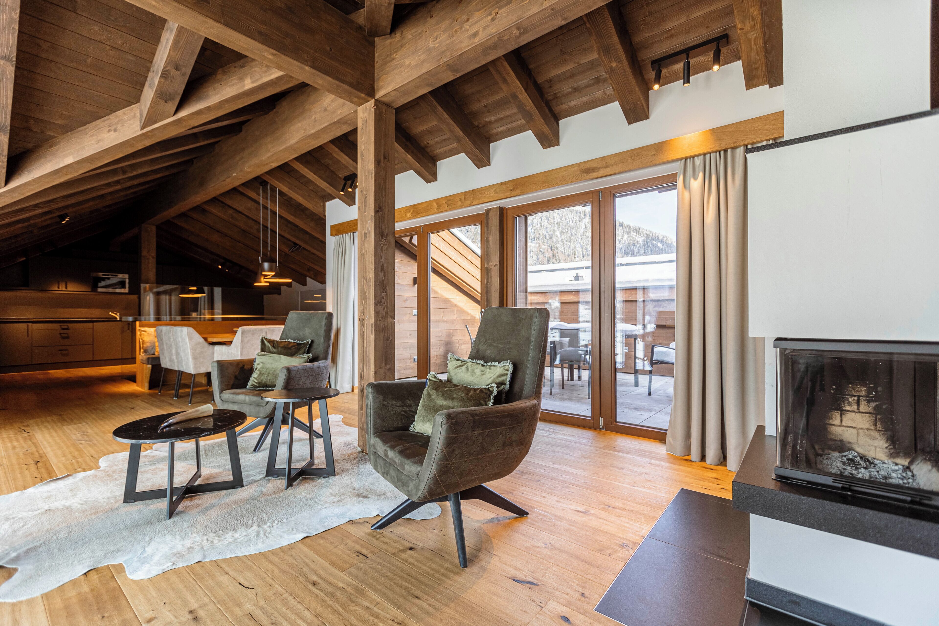 A spacious living area with wooden beams, a fireplace, and a large window overlooking a snowy landscape.