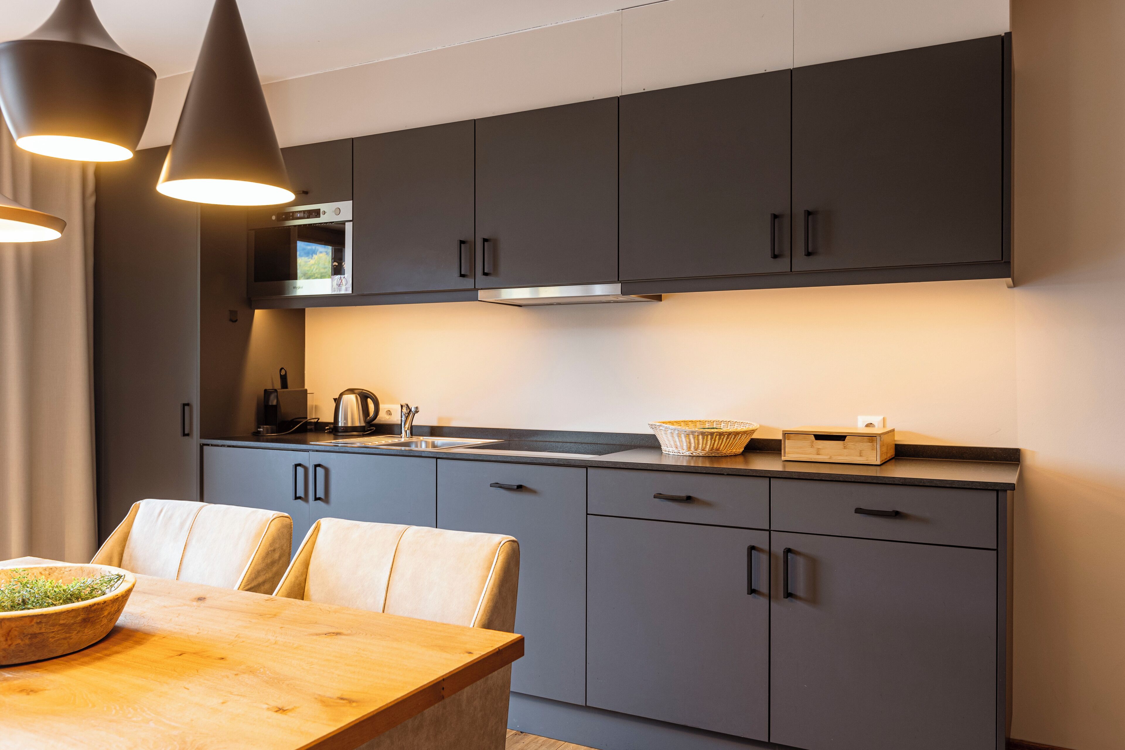 A modern kitchen with grey cabinets, a wooden dining table, and pendant lighting.
