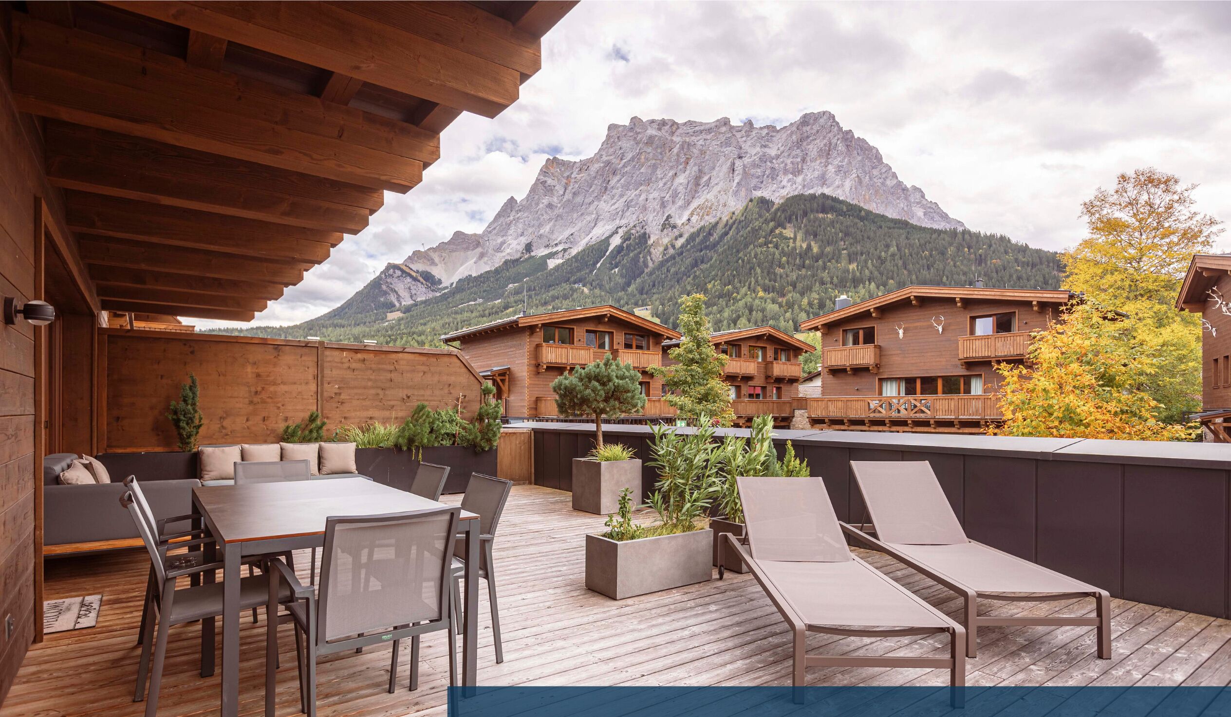 A wooden deck with a table and chairs, a wooden building with balconies, and a mountain in the background.