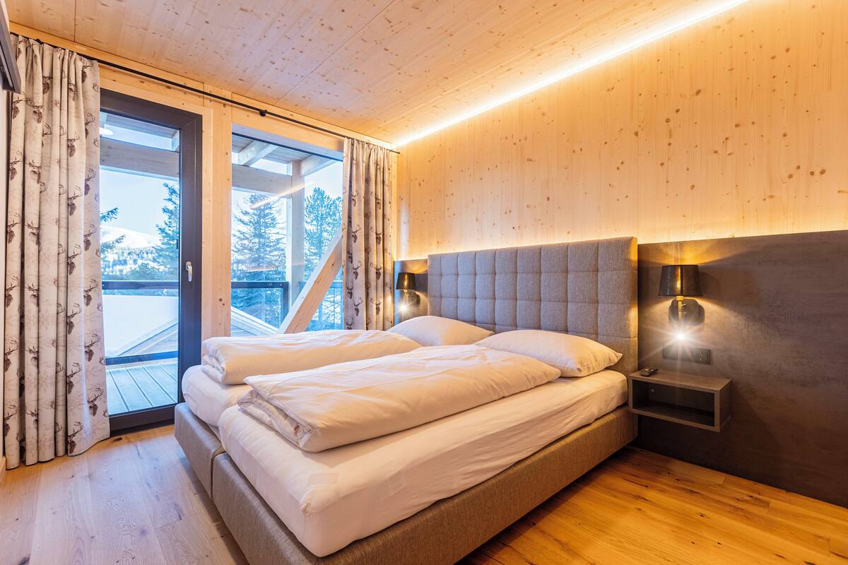 A modern bedroom with a large bed, wooden walls, and a view of a snowy landscape through a glass door.