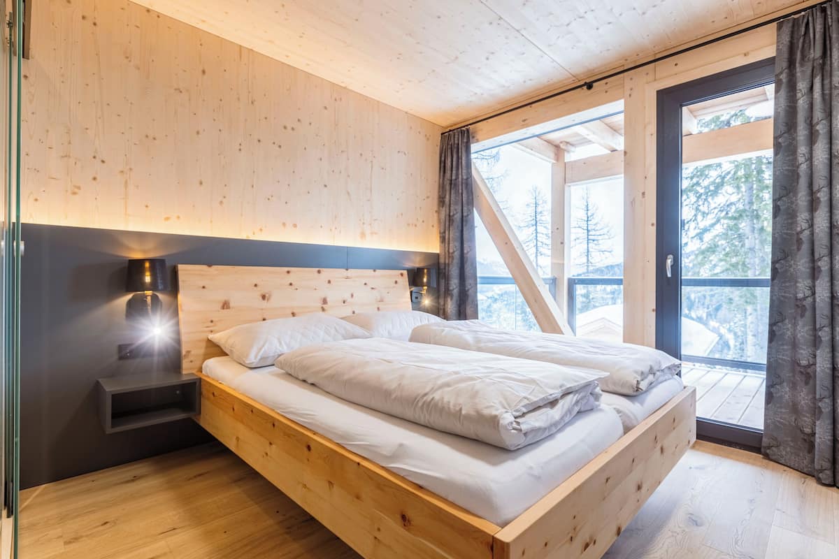A wooden bed with white bedding in a room with a sloped ceiling and large windows.