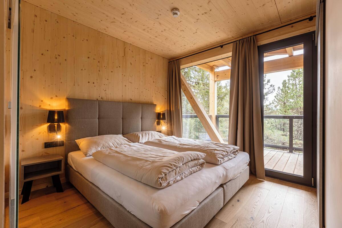 A bedroom with a wooden ceiling, a bed with beige bedding, two bedside lamps, and a large window with curtains.