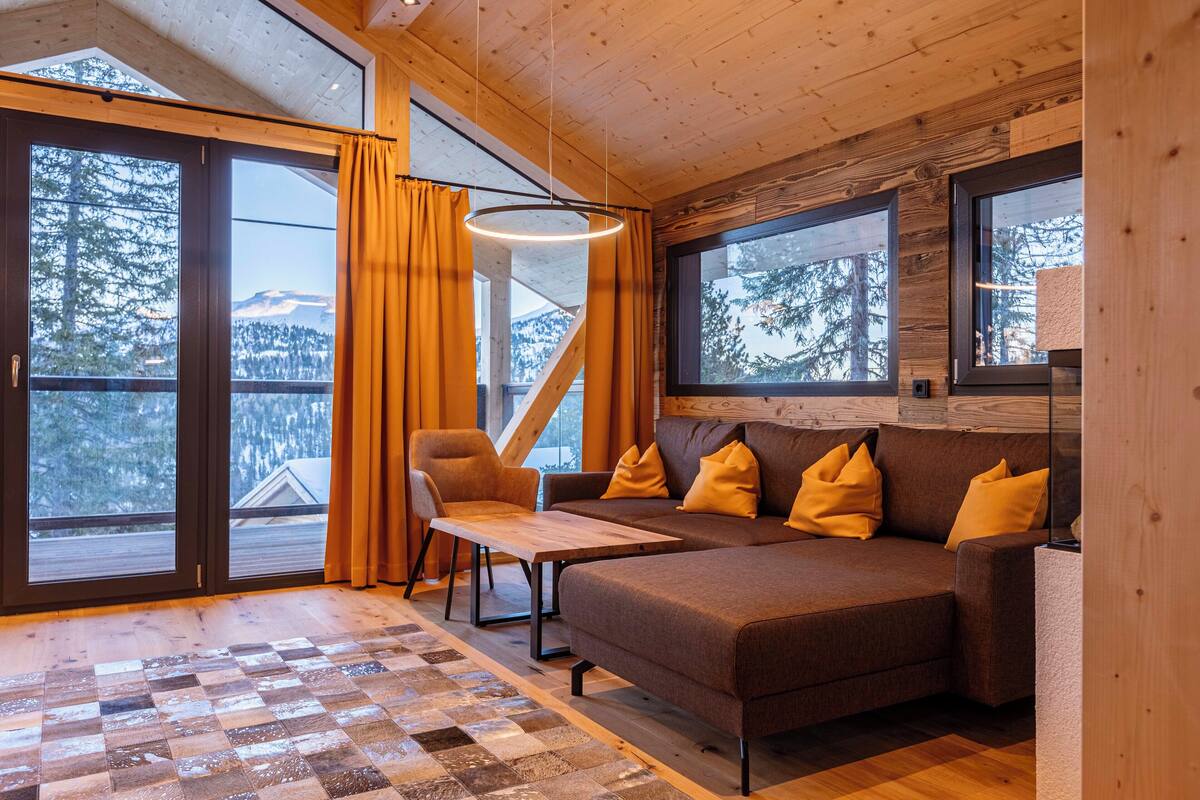 A cozy cabin interior with a wooden floor, a sofa with yellow cushions, a wooden table, and large windows offering a view of the forest and mountains.
