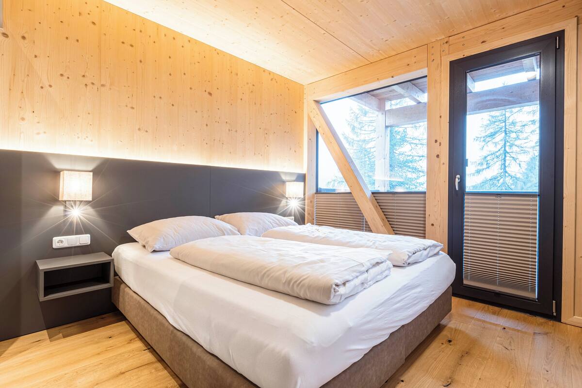 A modern bedroom with a wooden ceiling, a bed with white linens, a nightstand with a lamp, and a window with blinds.