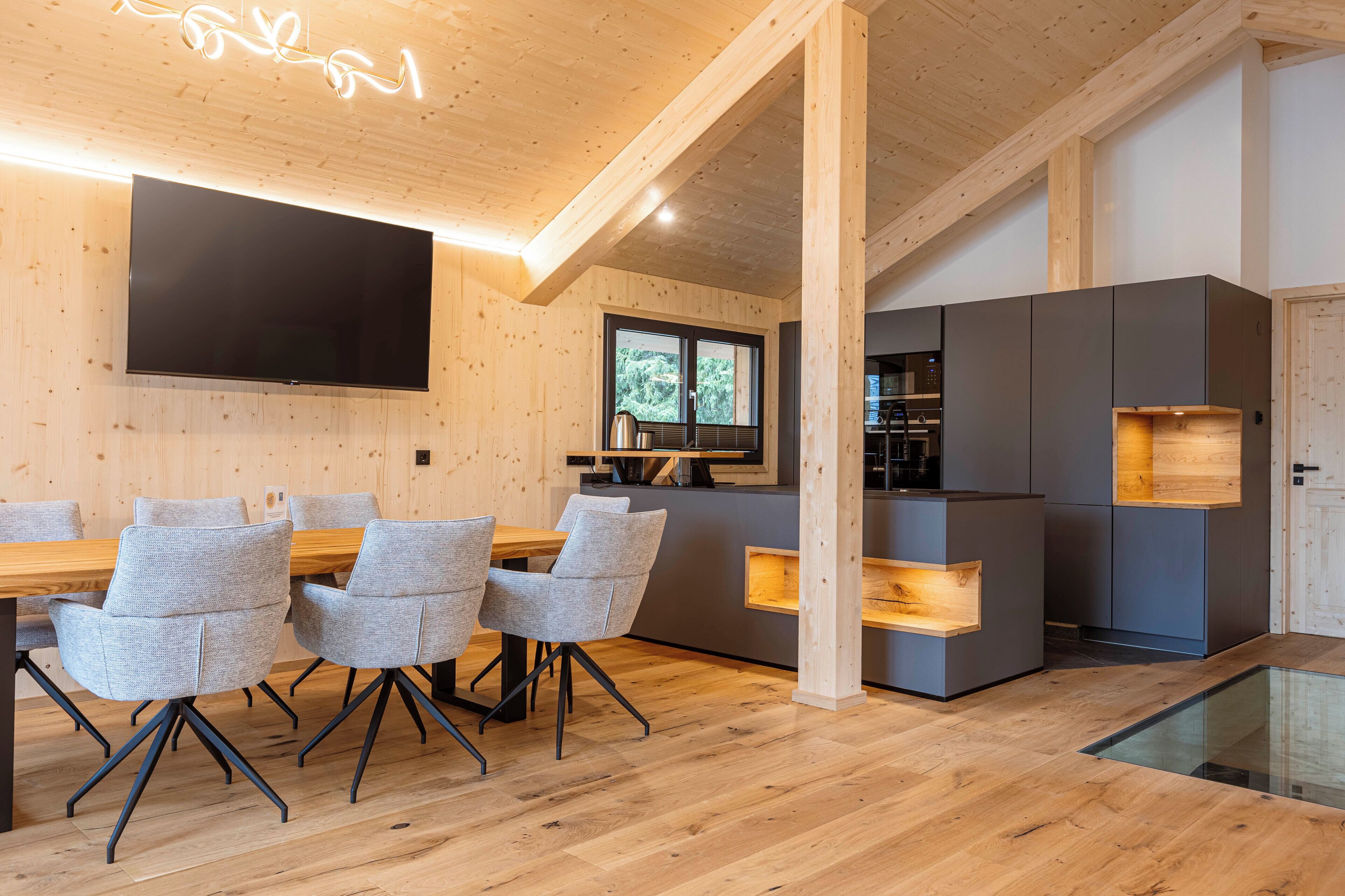 A modern interior with a wooden ceiling, a flat-screen TV, a dining area with grey chairs, and a kitchen with grey cabinets.