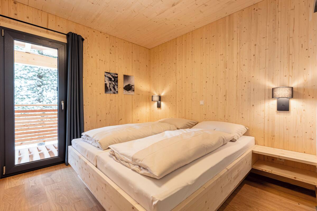 A wooden-paneled room with a bed, a window with curtains, and two wall-mounted lights.