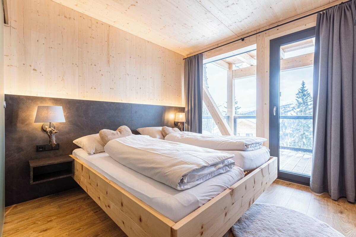 A wooden bedroom with a large bed, two bedside tables, and a view of a snowy landscape through a glass door.