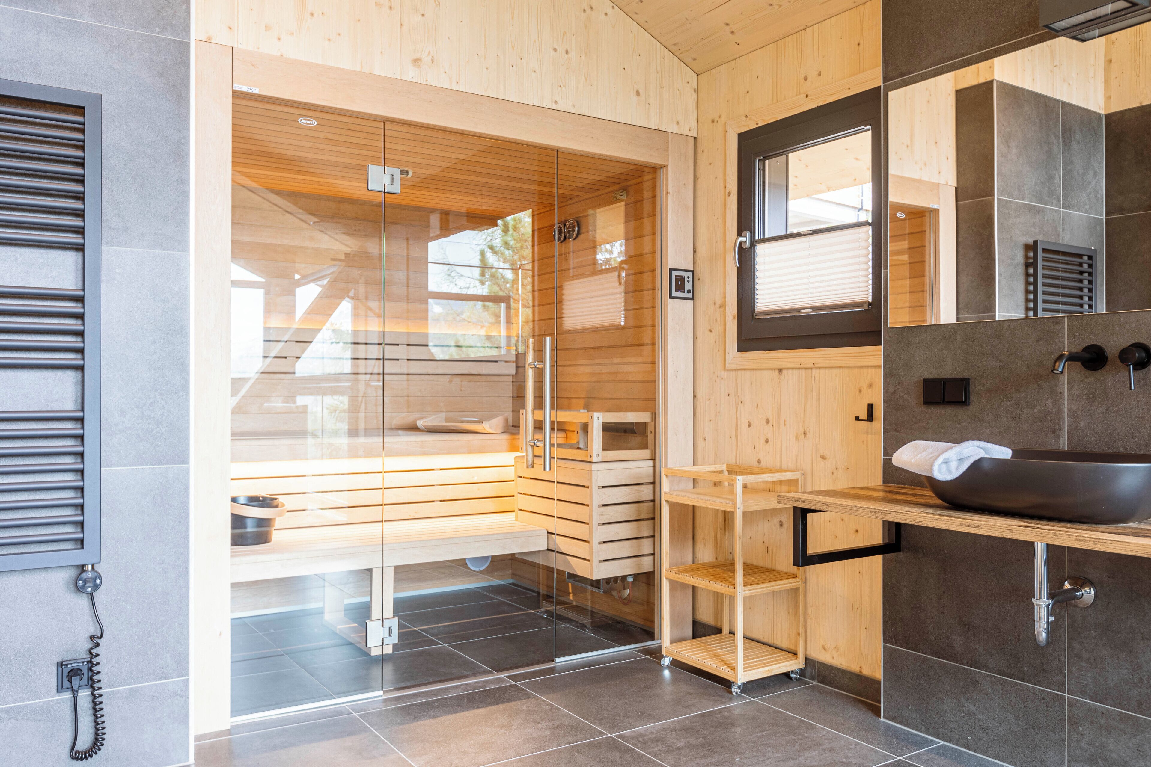 Un baño moderno con detalles de madera, una sauna con paredes de vidrio, un lavamanos grande de color negro y un espejo encima.