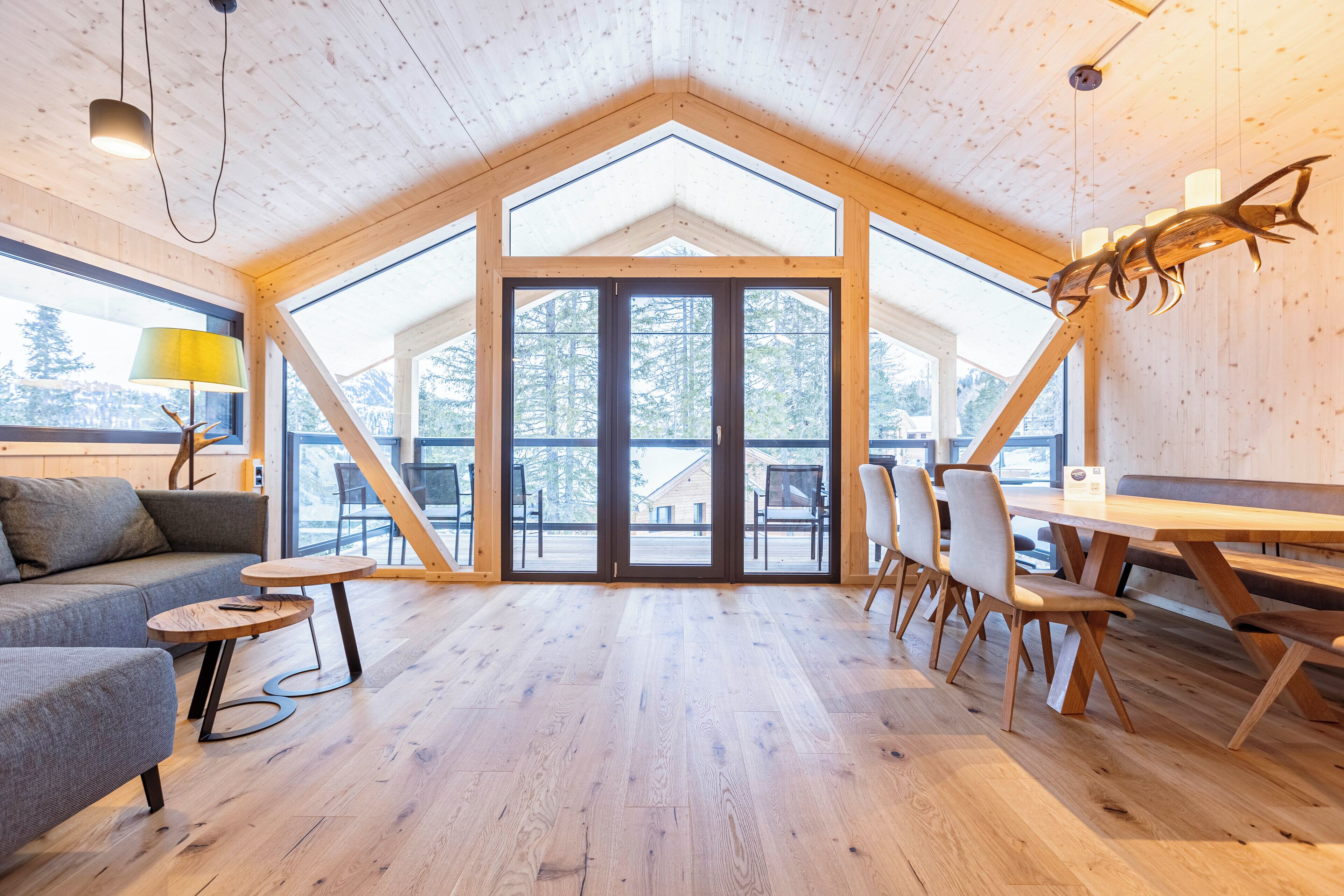 A spacious room with a high ceiling, wooden flooring, a large window, and a dining area with chairs and a table.