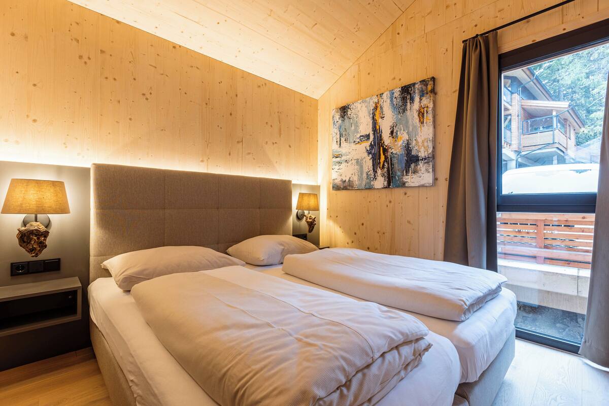A hotel room with a wooden ceiling, a bed with white bedding, two bedside lamps, a wall-mounted artwork, and a window with curtains.