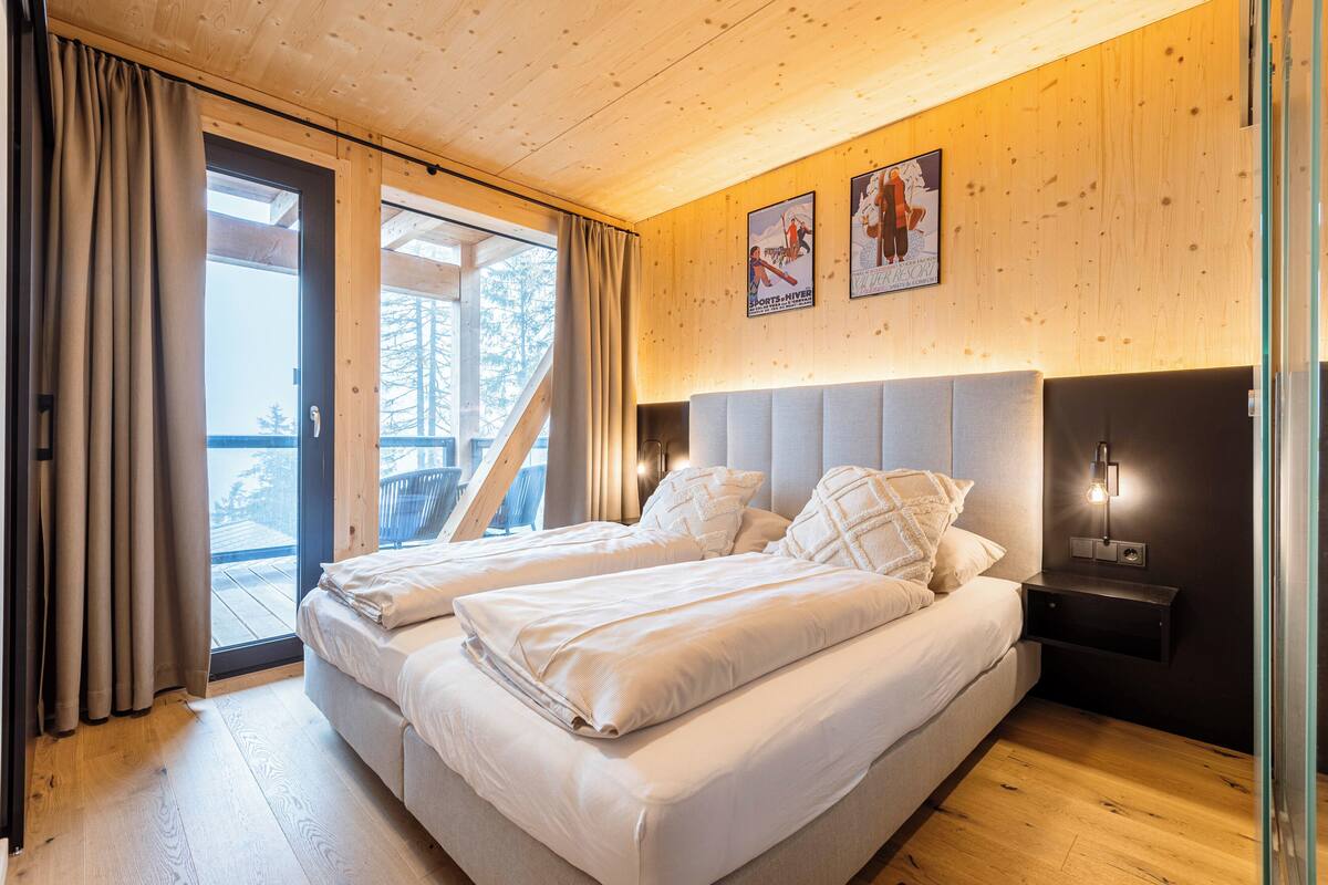 A bedroom with two beds, a wooden ceiling, a nightstand with a lamp, and a view of the outdoors through a glass door.