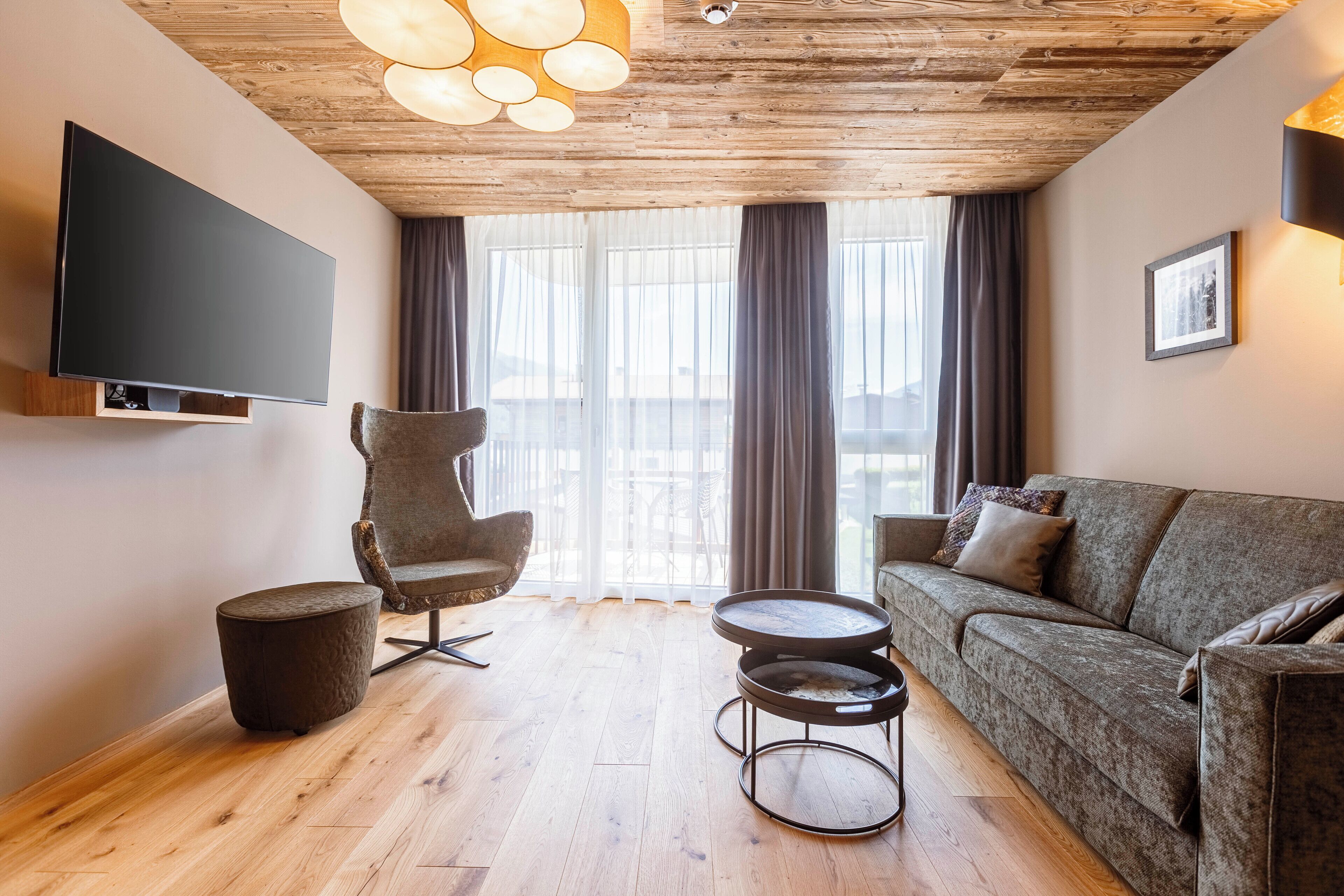A modern living room with a wooden ceiling, a flat-screen TV mounted on the wall, a grey sofa, a round coffee table, and a high-back chair.