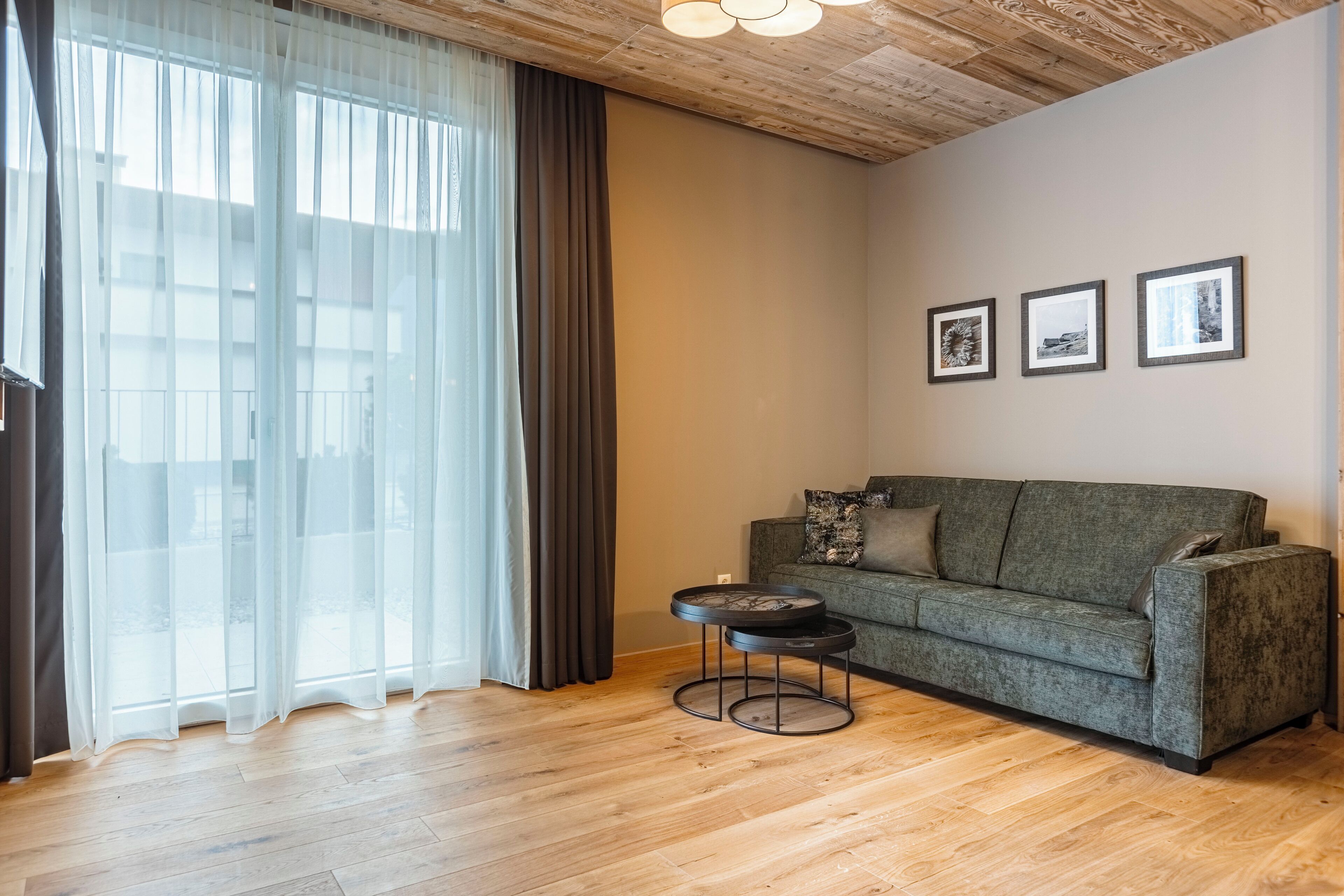 A living room with a wooden floor, a grey sofa, a round coffee table, and a large window with sheer curtains.