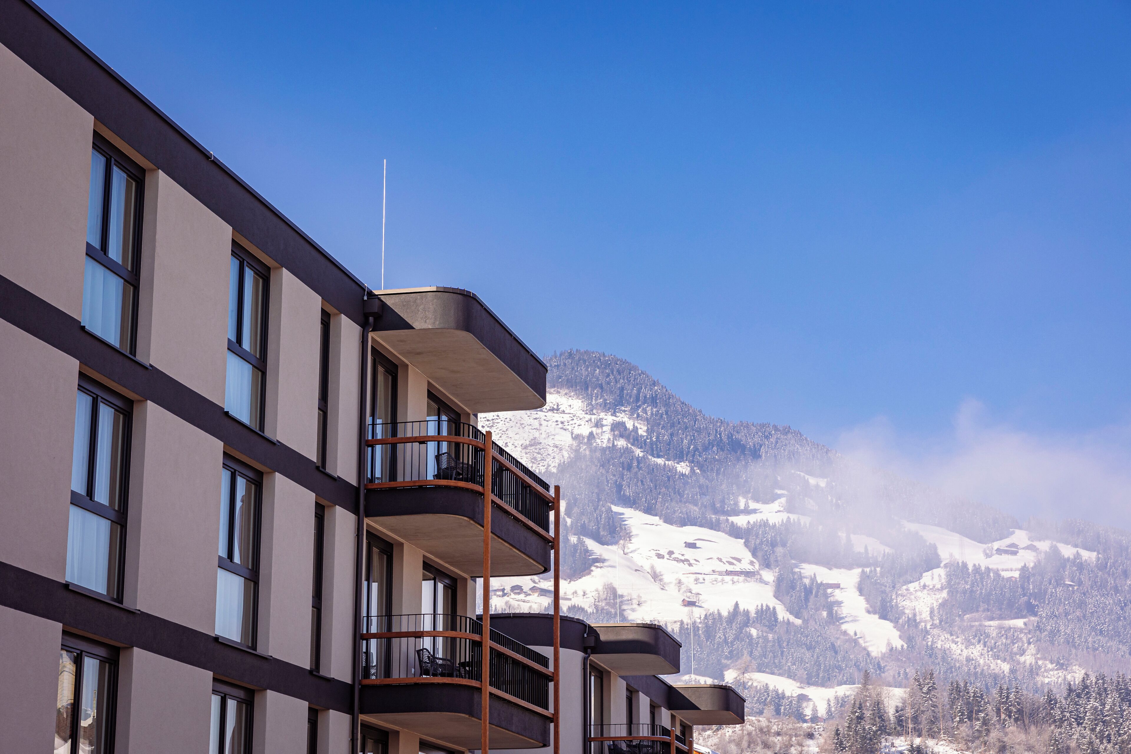 A multi-story hotel with balconies, a clear view of snow-covered mountains, and a flag on the roof.