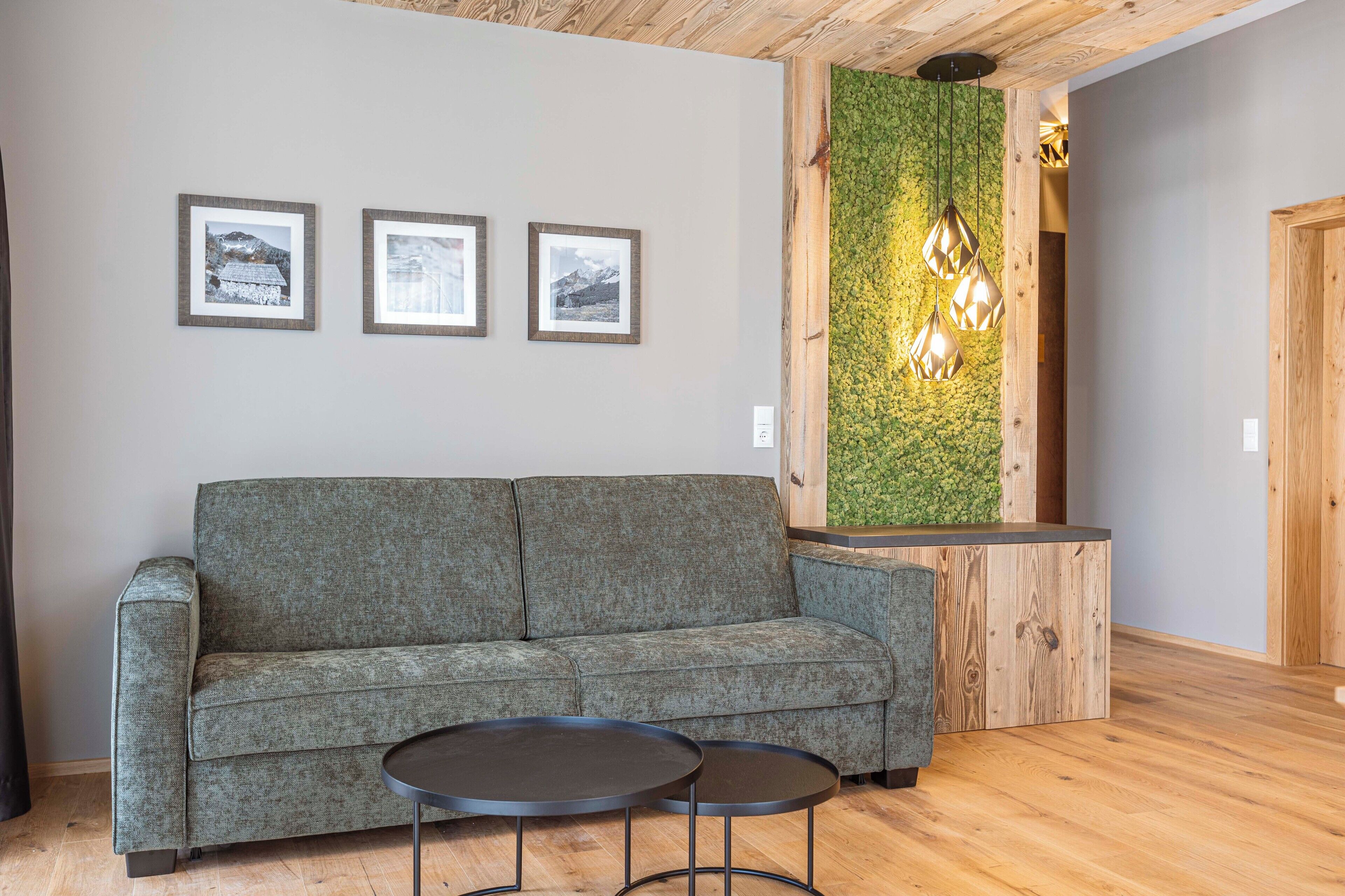 A modern lobby with a grey sofa, two round tables, framed pictures on the wall, and a green wall with hanging lights.