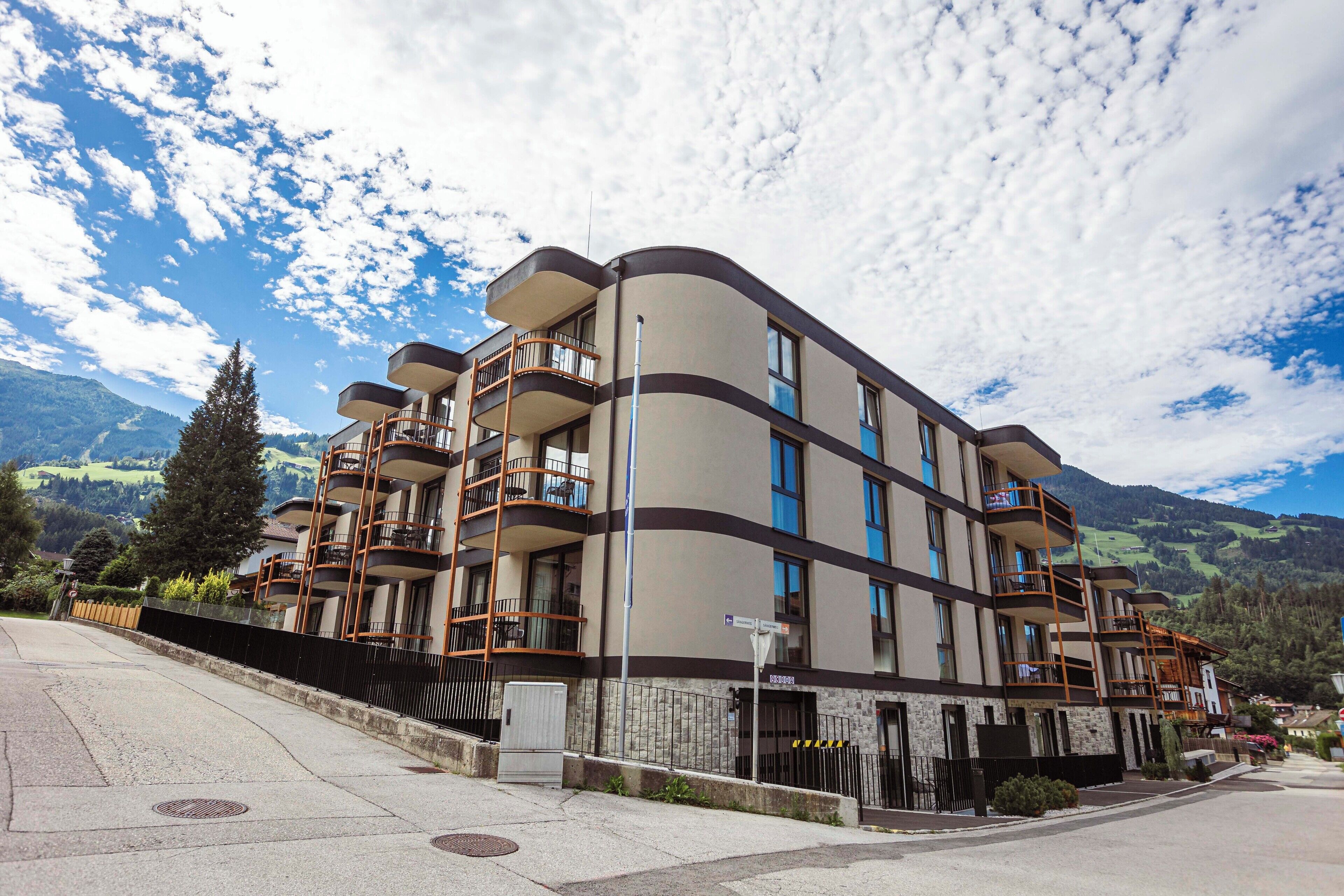 A multi-story hotel with balconies, a stone facade, and a view of mountains in the background.