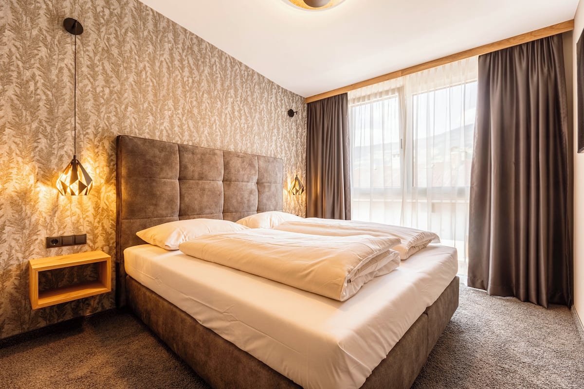 Un dormitorio con una cama grande, una mesita de noche de madera, una lámpara de pared y una ventana con cortinas.