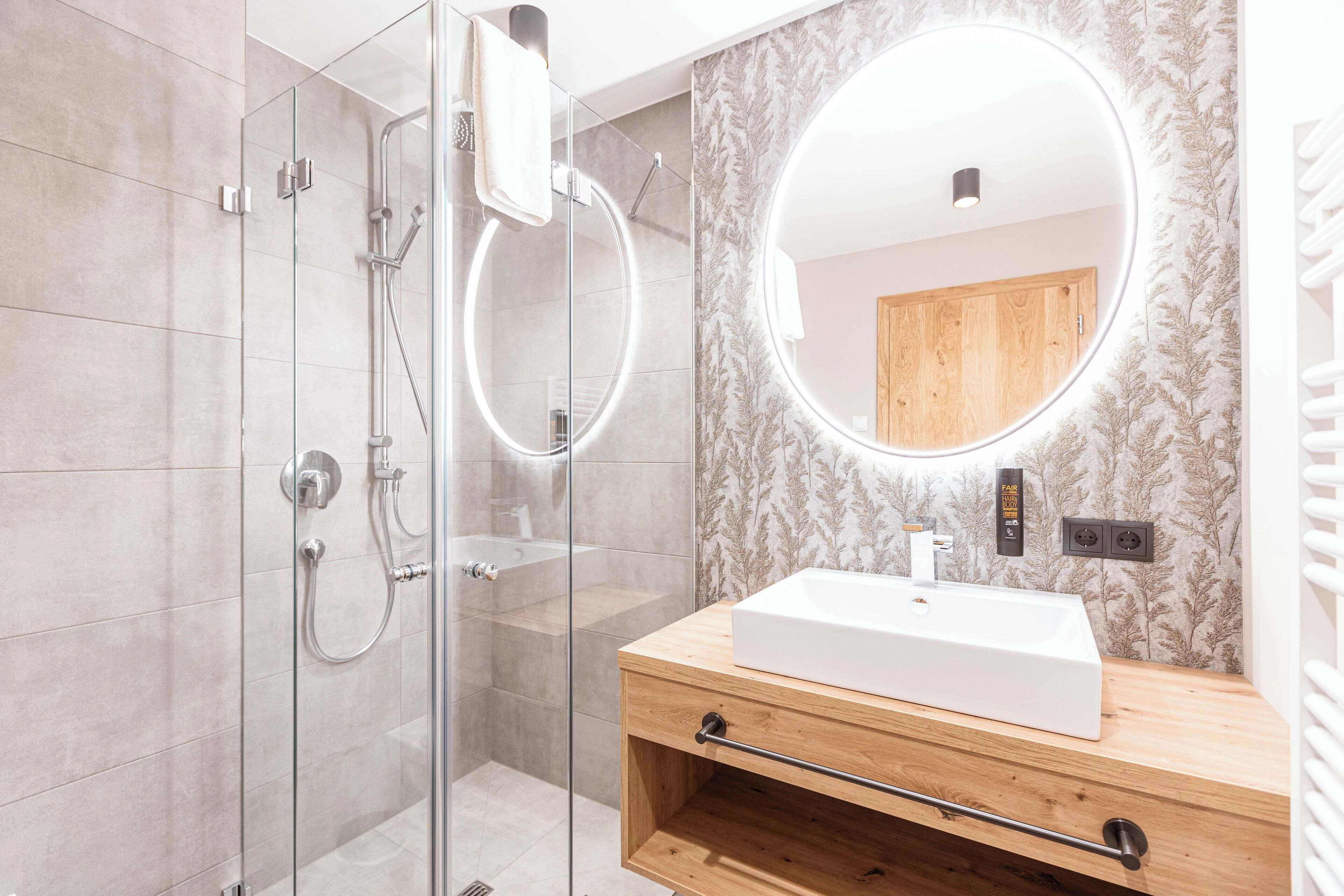 A modern bathroom with a glass-enclosed shower, a wooden vanity with a white sink, and a large mirror with integrated lighting.