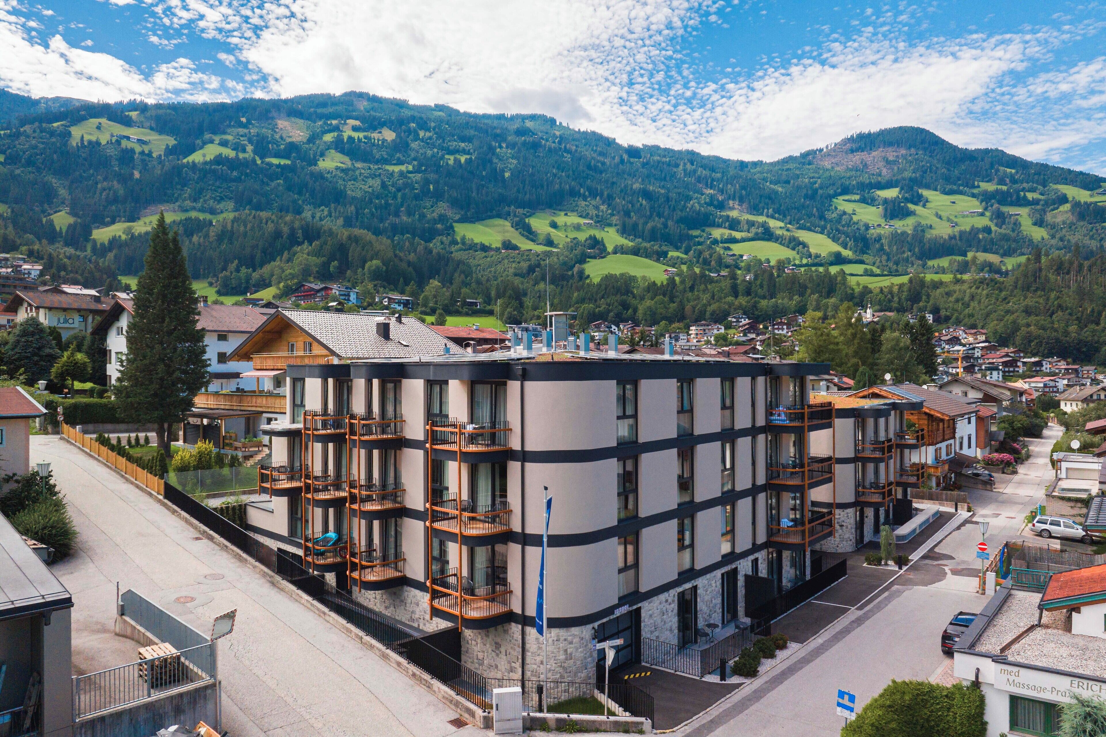 A multi-story hotel with balconies, situated in a mountainous area with visible greenery and houses in the background.