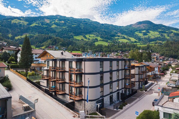 A multi-story hotel with balconies, situated in a mountainous area with visible greenery and houses in the background.