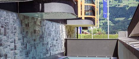 A swimming pool with a stone wall and wooden deck, a staircase, and a view of a mountainous landscape.