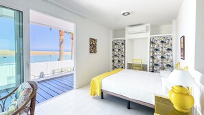 A bedroom with a bed, a chair, a bedside table with a lamp, a window with a view of the beach, and an air conditioning unit.