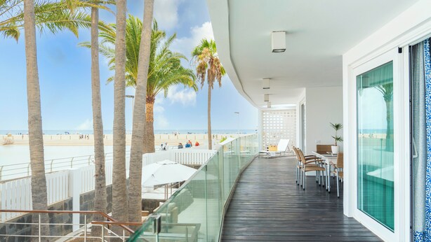 A balcony with a view of the beach, palm trees, and a dining area with chairs and tables.