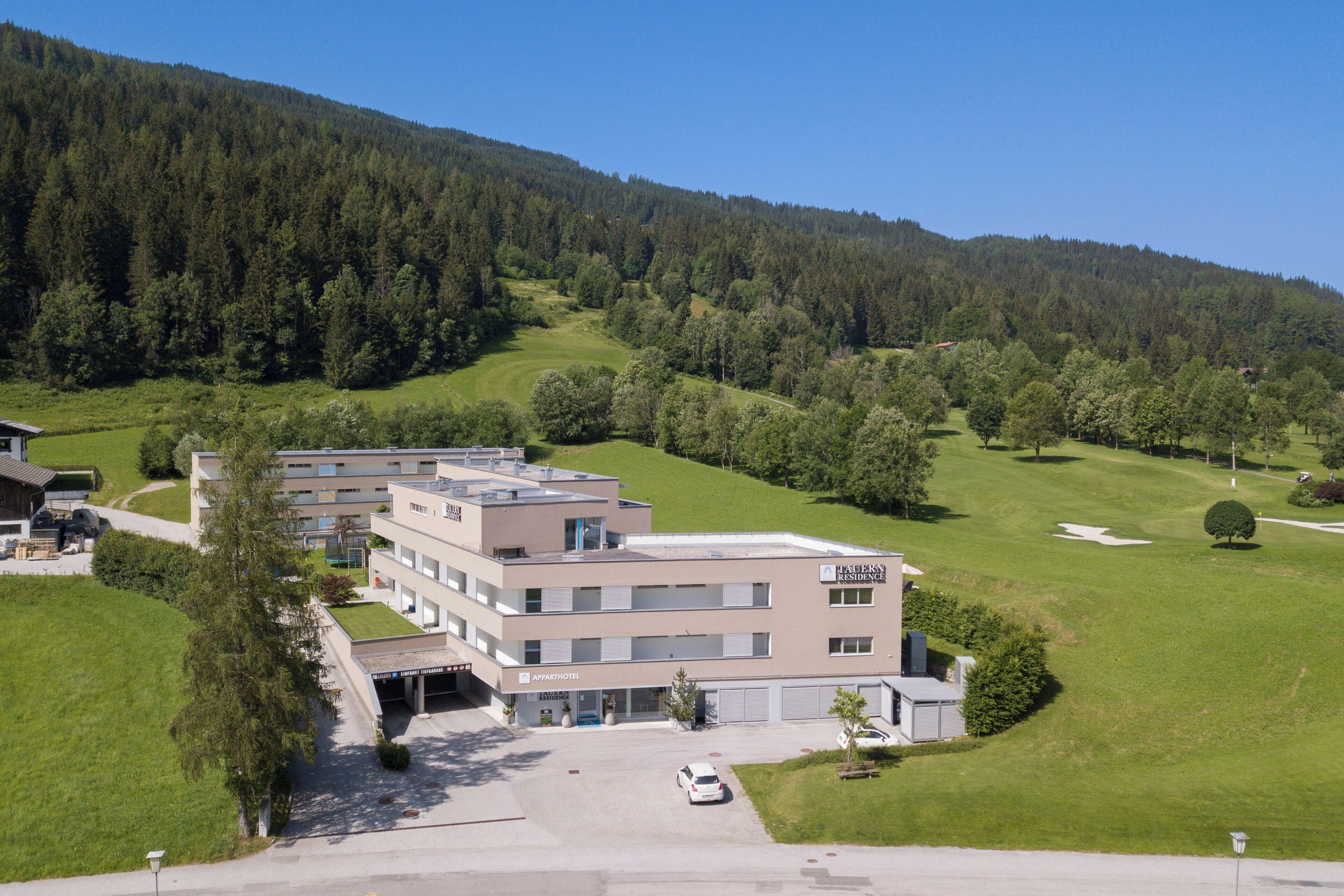 A multi-story hotel with a parking area, surrounded by lush greenery and a hilly landscape.