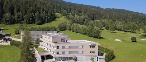 A multi-story hotel with a parking area, surrounded by lush greenery and a hilly landscape.