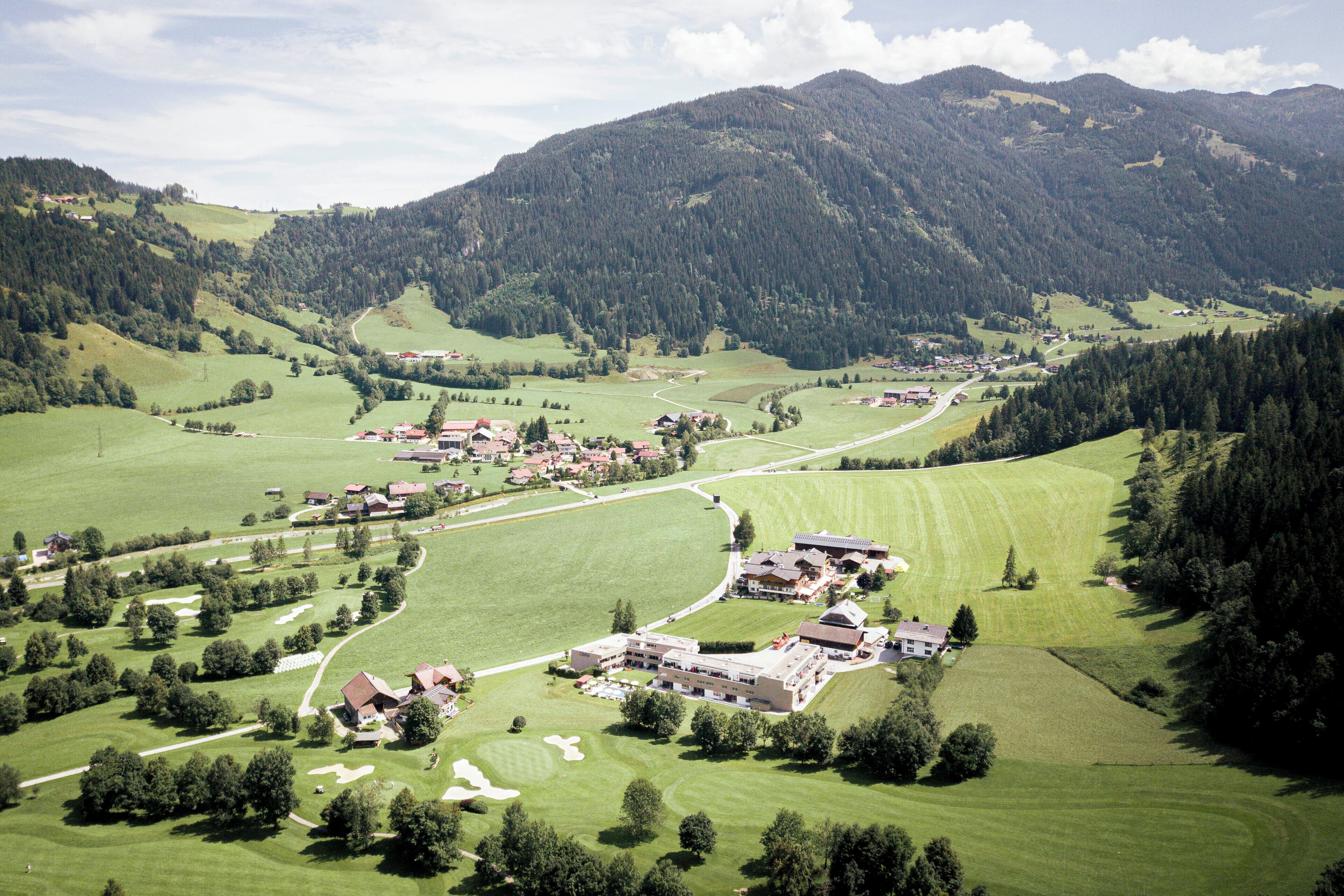 A rural village with houses and a large building, surrounded by green fields and mountains.