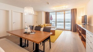 Habitación de hotel moderna con comedor, sofá con cojines, televisor y vista al mar.