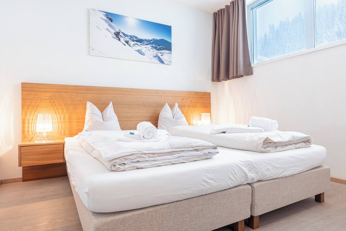Camera d'albergo con due letti, testiere in legno, biancheria bianca, un comodino con una lampada e un quadro raffigurante una montagna innevata.