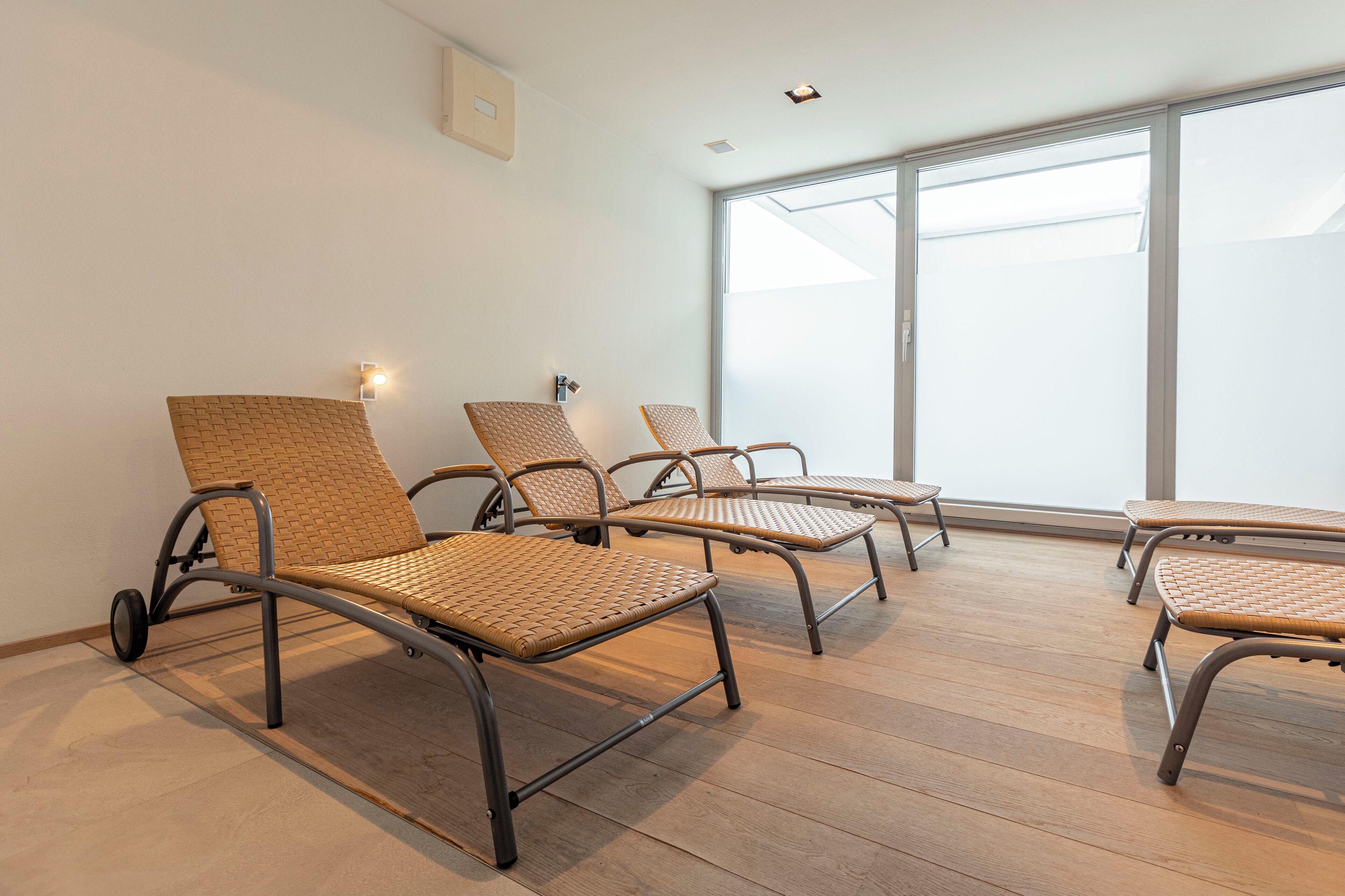 Una habitación con cuatro sillones de mimbre alineados, un ventanal que proporciona luz natural y una lámpara de techo.