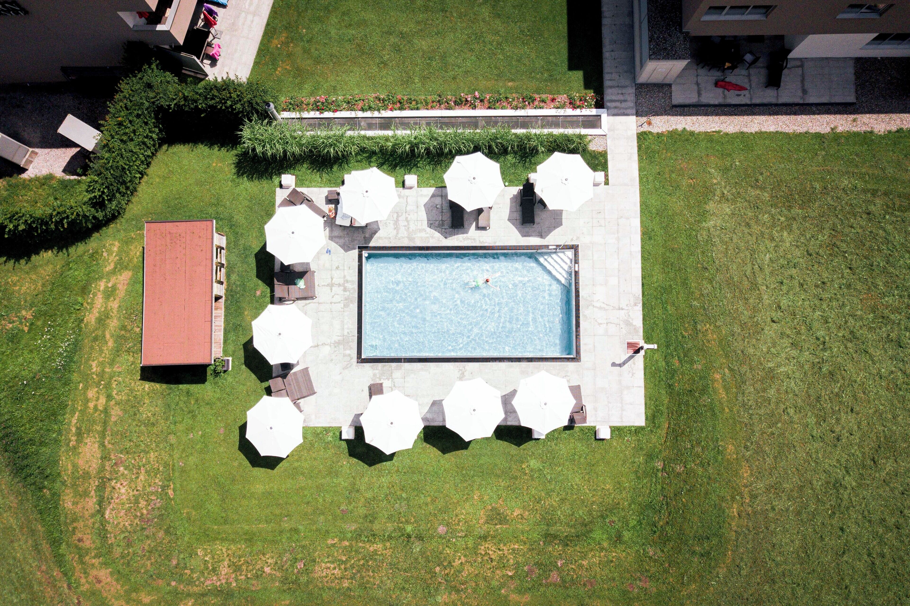 An aerial view of a swimming pool area with a rectangular pool, surrounded by white umbrellas and lounge chairs, and a small building to the left.