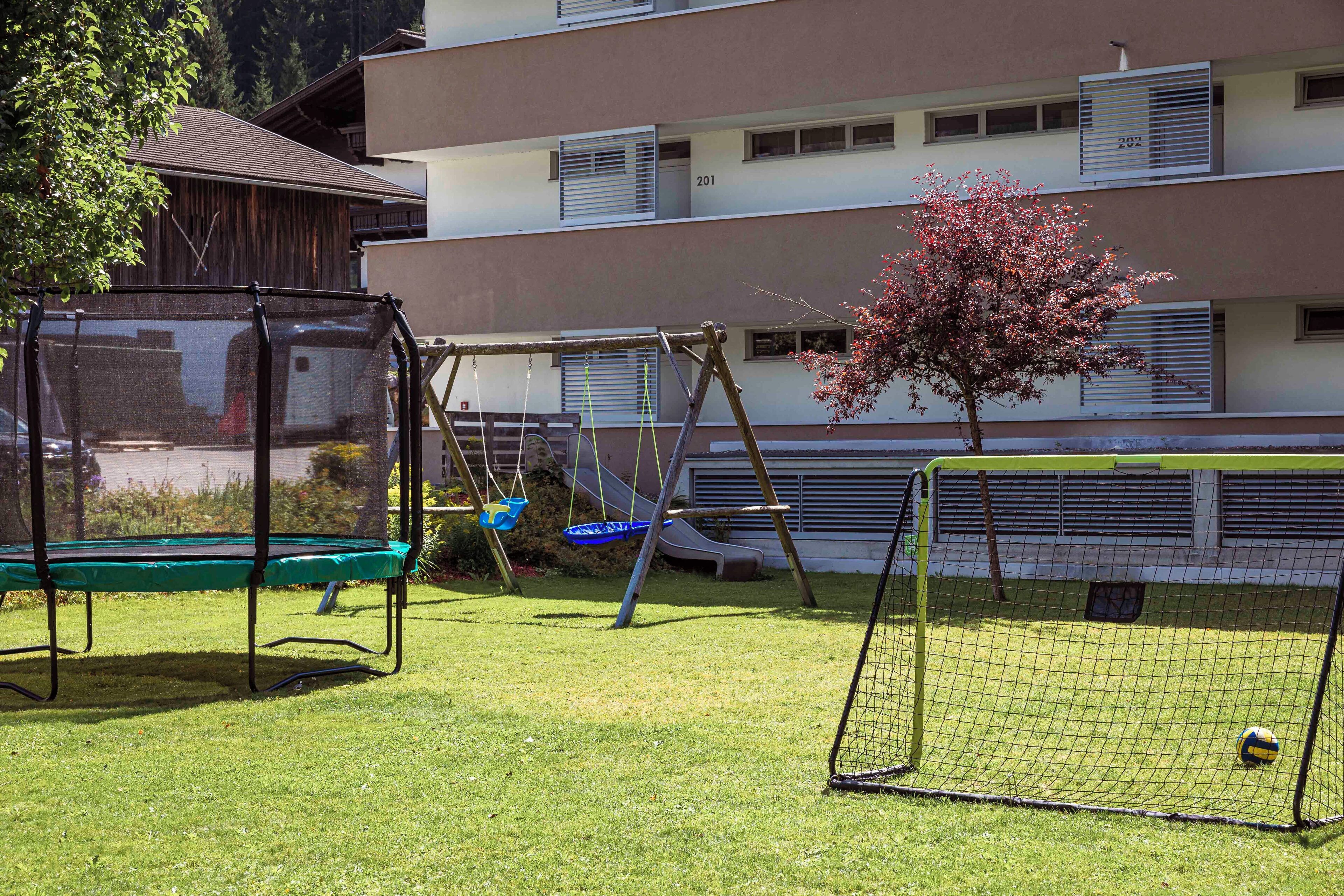 A playground with swings and a trampoline, a soccer goal, and a building with multiple windows in the background.