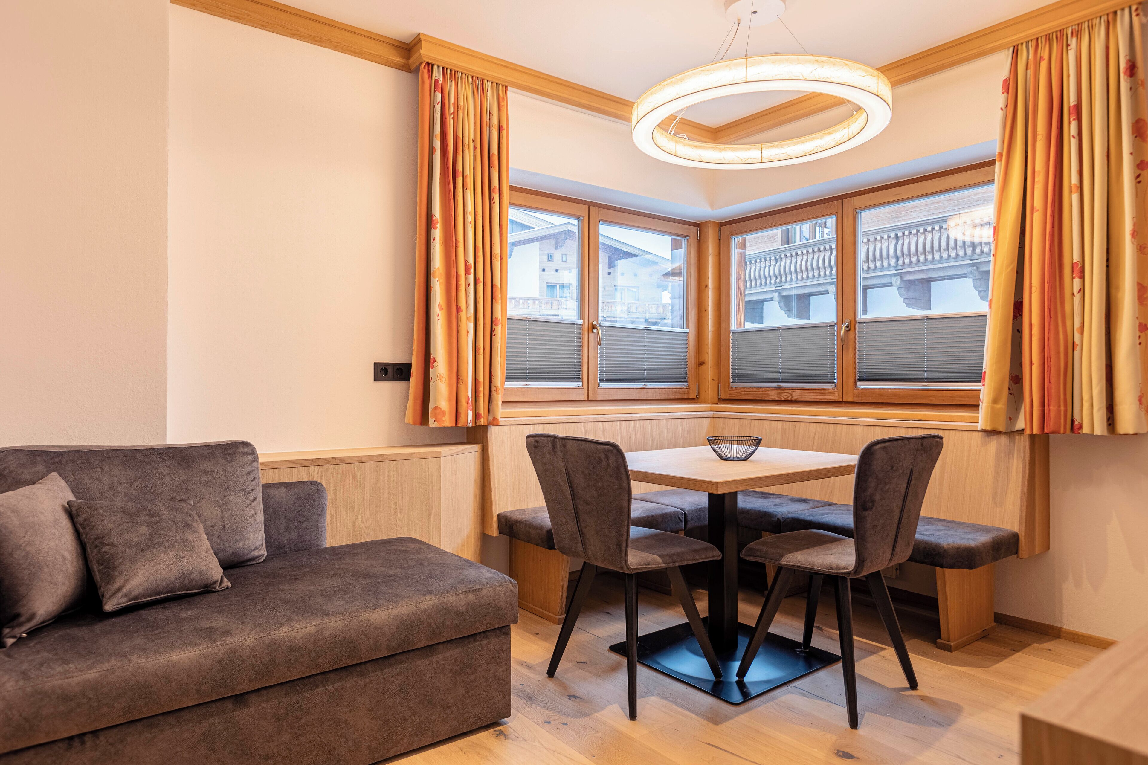 A modern living room with a sofa, a dining table, and chairs. There is a large window with curtains, and a circular light fixture above the table.