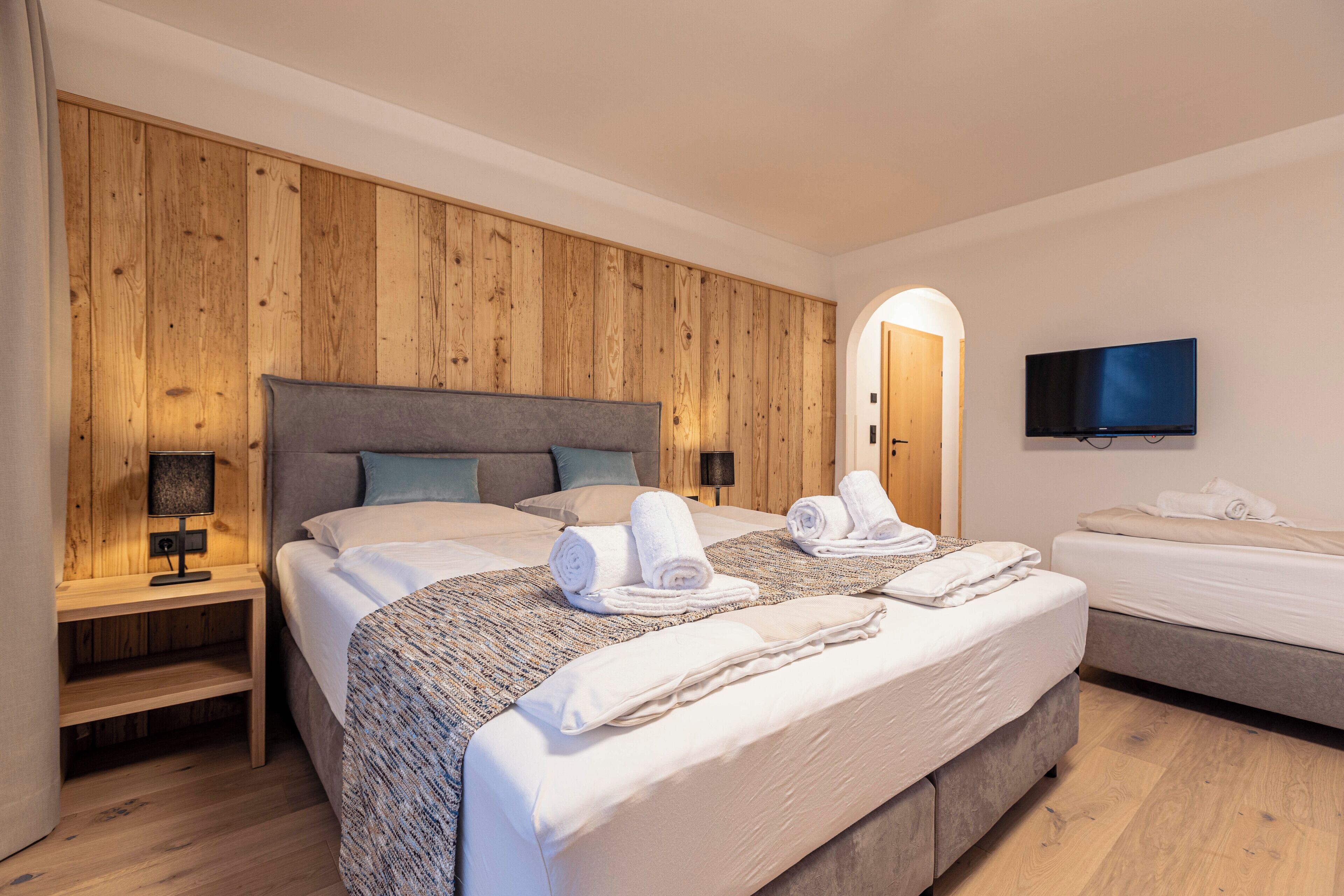 A hotel room with a large bed, a smaller bed, a TV mounted on the wall, and a wooden panel wall.